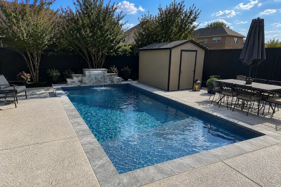 Vinyl inground pool with a raised stone waterfall wall, wood privacy fence, and patio dining set